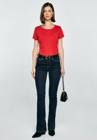 Červené tričko s krátkým rukávem a kulatým výstřihem, se zlatým logem; zkombinováno s tmavě modrými úzkými džíny, černým páskem a malou černou kabelkou.