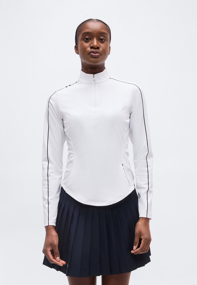 Γυναίκα που φοράει λευκή μακρυμάνικη ζακέτα με φερμουάρ και μαύρα περιγράμματα, και μπλε ναυτικό πλισέ φούστα, στέκεται μπροστά σε έναν απλό φόντο.