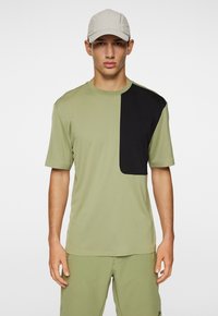 Oliivinvihreä t-paita, jossa on musta kyynärpäälappu vasemmalla puolella, lyhyet hihat, pyöreä kaula-aukko, valmistettu kevyestä kankaasta. Yhdistetty oliivinvihreisiin shortseihin.