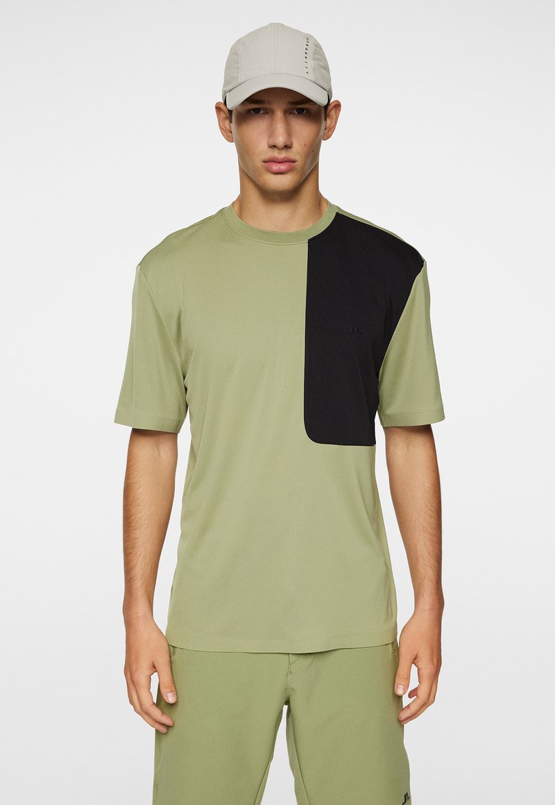 Oliivinvihreä t-paita, jossa on musta kyynärpäälappu vasemmalla puolella, lyhyet hihat, pyöreä kaula-aukko, valmistettu kevyestä kankaasta. Yhdistetty oliivinvihreisiin shortseihin.