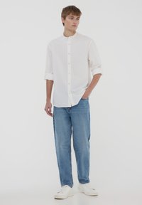 Camicetta bianca con colletto eretto, maniche arrotolate, abbinata a jeans azzurri a vestibilità rilassata e sneakers bianche. Design semplice e pulito.