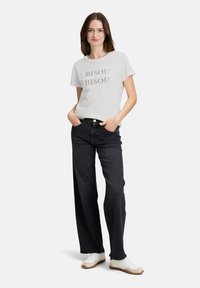 Hvid t-shirt med "BISOU BISOU" trykt i grå, kombineret med højhalsede, vide sorte jeans og hvide sneakers. Model poserer med hænderne i lommerne.