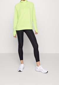 Helles grünes Langarm-Sportoberteil mit einem Halbreißverschluss, kombiniert mit schwarzen Leggings mit Seitentaschen und weißen Turnschuhen.