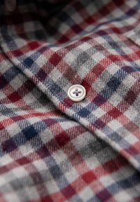 Camicia di flanella con un pattern a quadri grigio, rosso e blu, caratterizzata da un bottone bianco con design a quattro fori e una texture morbida.