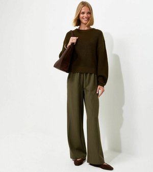Femme souriante, portant un pull vert foncé, un pantalon large à rayures, des ballerines marron et tenant un sac à bandoulière marron sur fond blanc.