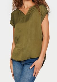 Blusa de manga corta en verde oliva con escote en V, que presenta un sutil patrón de puntos y una tela suave y ligera. Con dobladillo curvo y ajuste relajado.