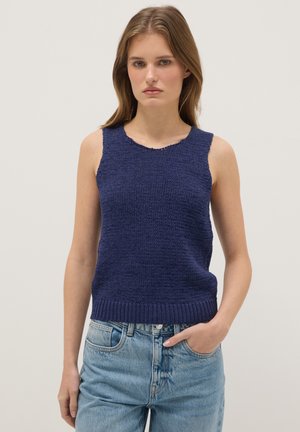 Mujer con un top de punto azul marino sin mangas y vaqueros azul claro, de pie con una mano en el bolsillo contra un fondo claro sencillo.