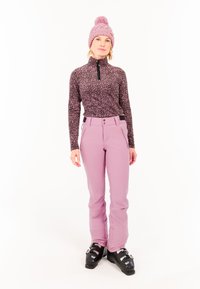 Fleece top s čiernym a ružovým leopardím vzorom, vysokým golierom a zipsom. Svetloružové lyžiarske nohavice a čierne lyžiarske topánky s kovovými sponami.