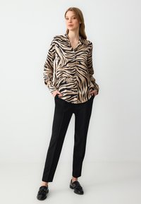 Beige en zwarte zebra print blouse met lange mouwen en knopen, gecombineerd met zwarte slim-fit broeken en zwarte loafers op een effen achtergrond.