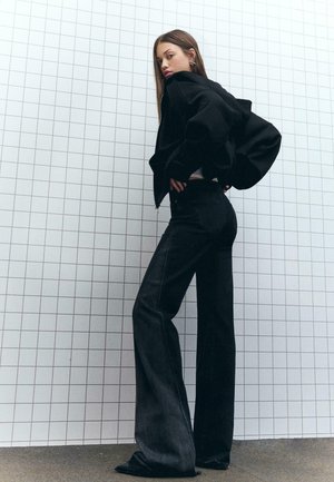 Black cropped jacket with voluminous sleeves, paired with high-waisted dark denim flared jeans. Model stands against a grid-patterned wall.
