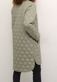 Kviltad lång jacka i dämpad grön, med diamantmönster, sidfickor och en lätt böjd nederkant. Strukturerad tyg.