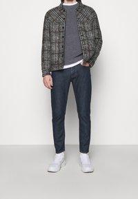 Chaqueta de cuadros en tonos marrones y grises sobre un suéter de punto gris, combinada con jeans azul oscuro y zapatillas blancas.