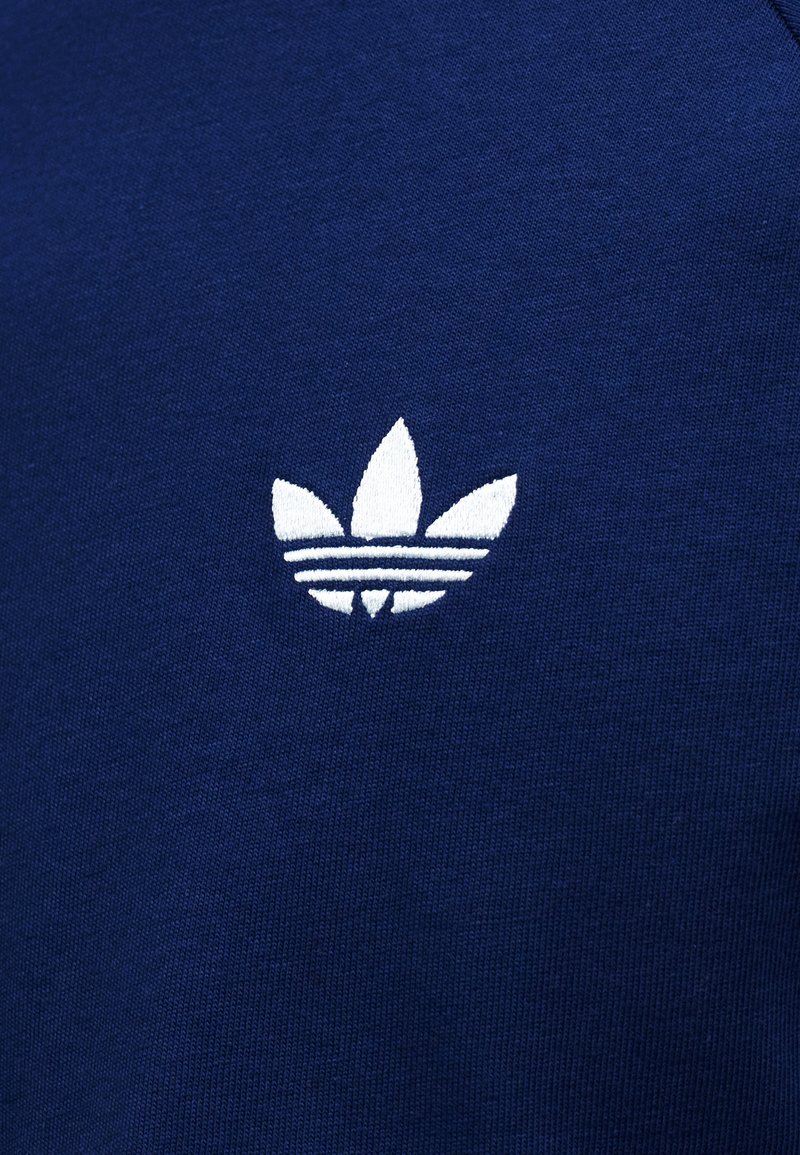 Logo Adidas trefoil ricamato bianco su uno sfondo di tessuto blu scuro con texture, centrato nella cornice.