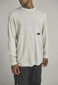 Langärmliges, hellgraues Shirt aus weichem Material, mit einer horizontalen Naht und einem kleinen schwarzen Markenlabel auf der Vorderseite.