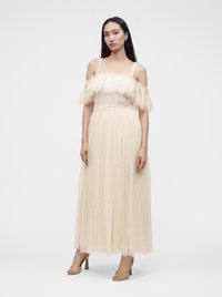 Femme aux longs cheveux foncés portant une robe en dentelle crème à épaules dénudées et des talons ouverts dorés, debout devant un fond blanc uni.