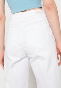 Pantaloni in denim bianco a vita alta con una vestibilità comoda, dotati di due tasche posteriori, dettagli cuciti e una texture liscia.