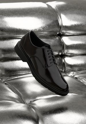 Scarpa elegante nera lucida con lacci, posata su una superficie argento imbottita e lucida in pelle.