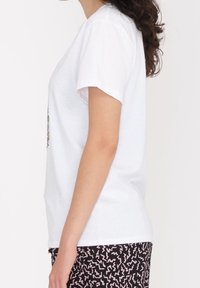 Hvid bomulds-t-shirt med korte ærmer og rund hals, med et farverigt blomstermønster på forsiden. Sammen med mønstrede sorte bukser.