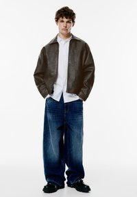 Veste en simili cuir marron avec un col, chemise blanche boutonnée et jeans en denim bleu surdimensionnés. Chaussures noires chunky avec des accents métalliques.