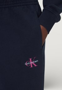 Granatowe spodnie dresowe z miękkiego materiału mają elastyczny pas i mankiety. Na lewym udzie haftowane jest białe i różowe logo Calvin Klein.
