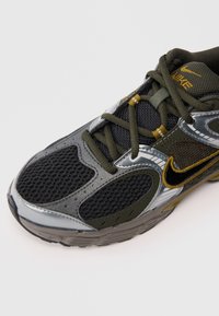 Sportskih cipela s crnom mrežicom na gornjem dijelu, metalno srebrnim i maslinasto zelenim akcentima te žutim detaljima na vezicama. Istaknuti swoosh logo.