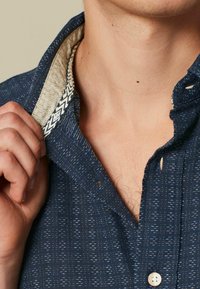 Camicia azzurra con bottoni, realizzata in un tessuto texturizzato, presenta un colletto foderato di beige e nero. Il colletto è leggermente sbottonato, rivelando parte del collo.