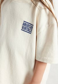 T-shirt en coton couleur crème avec une coupe ample, présentant un logo bleu "AMERICAN VINTAGE" sur le côté gauche. Manches courtes avec une texture subtile.