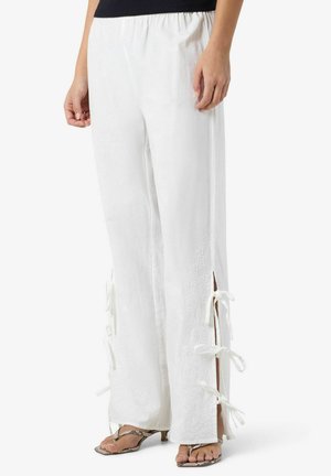 Žena nosiaca biele široké nohavice s bočnými rázporkami a zviazanými mašličkami, skombinované so sandálmi na podpätku so vzorom hadej kože.