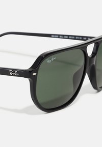 Ochelari de soare din plastic negru cu lentile verzi, având sigla Ray-Ban. Design cu ramă pătrată și accente din metal pe tâmple.