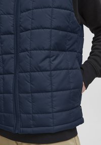 Marinblå vadderad puffer-väst med dragkedja och sidofickor, med en slät nylonutsida och kontrasterande mörka ärmar.