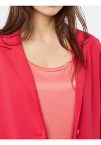 Blazer rouge vif en tissu léger, avec un col cranté et une fermeture à un seul bouton, superposé à un chemisier en satin rose.