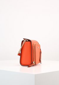 Liten orange tvärs över kroppen-väska med guldblixtlås och kedjesnodd, uppställd på vit yta mot en enkel vit bakgrund.