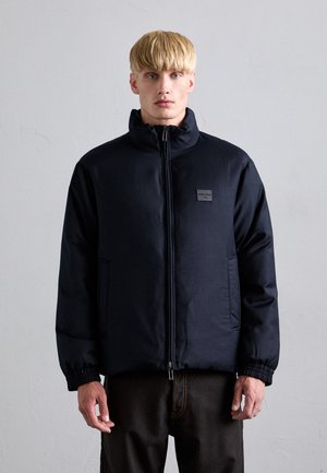 Marineblau gesteppte Jacke mit hohem Kragen, Reißverschluss, Seitentaschen und elastischen Bündchen. Verfügt über ein kleines Logopatch auf der Brust.