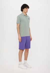 Zelené polo tričko s krátkymi rukávmi, s malým logom, v kombinácii s fialovými šortkami. Biele tenisky dopĺňajú celkový outfit.