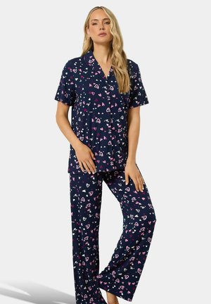 Femme aux longs cheveux blonds debout, portant un ensemble pyjama bleu marine à manches courtes, orné de motifs en forme de cœurs roses et blancs.