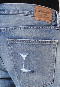 Jeans de mezclilla azul claro con un bolsillo trasero que presenta un pequeño desgarro desgastado y una etiqueta de cuero marrón de la marca Abercrombie & Fitch encima.