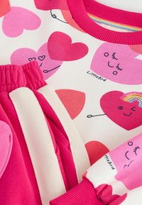 Set de îmbrăcăminte pentru copii, constând dintr-un tricou alb cu modele de inimioare roz și pantaloni scurți roz. Tricoul are manșete rib-knit; pantalonii scurți au dungi pe lateral.