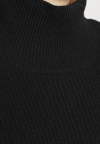 Gros plan sur un pull à col roulé en maille côtelée noire porté par une personne, mettant l'accent sur la texture et le design du col.