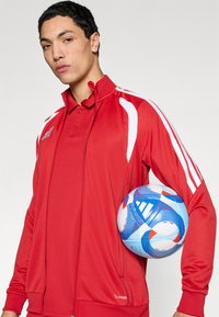 Κόκκινο αθλητικό μπουφάν με λευκές ρίγες, φερμουάρ μπροστά και γιακά τύπου πόλο, συνοδευόμενο από μια μπλε και λευκή μπάλα ποδοσφαίρου με σχέδιο.