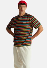 Proužkované tričko s krátkým rukávem s horizontálními pruhy v červené, modré, žluté a černé barvě, s logem "Stranger Things" na hrudi.