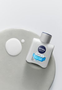 Nivea Men Sensitive Cool After Shave Balsam in einer klaren rechteckigen Flasche mit einem grauen Deckel, auf einem glatten grauen Cremeuntergrund platziert.