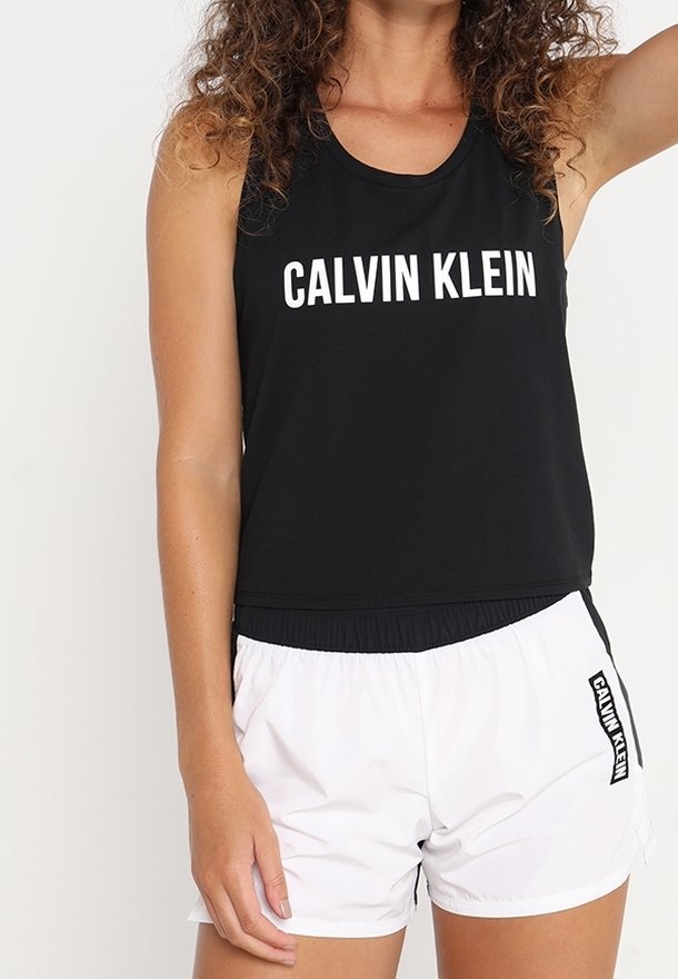 Sort ærmeløs top med "CALVIN KLEIN" i hvid tekst; kombineret med hvide shorts med en sort accentstribe og elastisk talje.