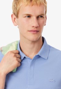 Jasnoniebieska koszulka polo z klasycznym kołnierzykiem i subtelnym logo Lacoste na piersi, noszona z wzorzystym ręcznikiem zarzuconym na jedno ramię.