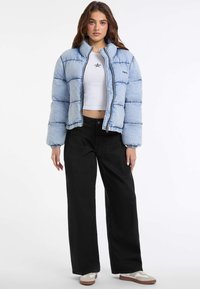 Lichtblauwe pufferjack met een hoge kraag, ritssluiting en gewatteerde textuur, gecombineerd met zwarte wijde broek en witte sneakers.