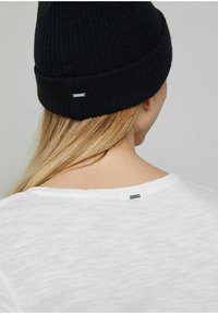 Černá žebrovaná pletená čepice s malým stříbrným akcentem, nošená s obyčejným bílým tričkem, zobrazená ze zadní strany s vlasy.