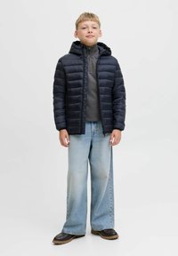 Veste matelassée bleu marine avec capuche, associée �à un haut gris à col montant, un large jean bleu clair et des chaussures à semelle marron. Tissu lisse, coupe décontractée.