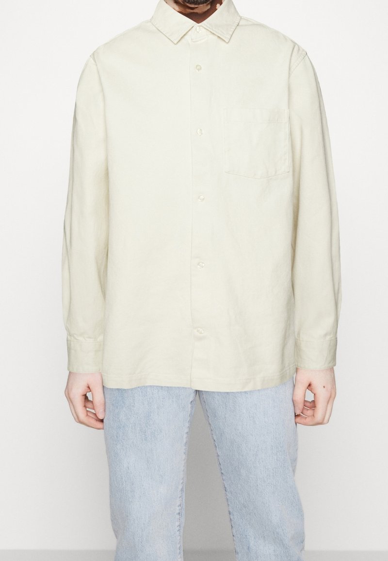 Homme portant une chemise crème à boutons à manches longues avec une poche poitrine et un jean en denim bleu clair, debout devant un fond blanc.