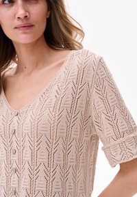 Jasno beżowy sweter z dzianiny, z dekoltem w kształcie litery V, krótkimi zrolowanymi rękawami i geometrycznym wzorem z koronki. Posiada cztery małe guziki z przodu.