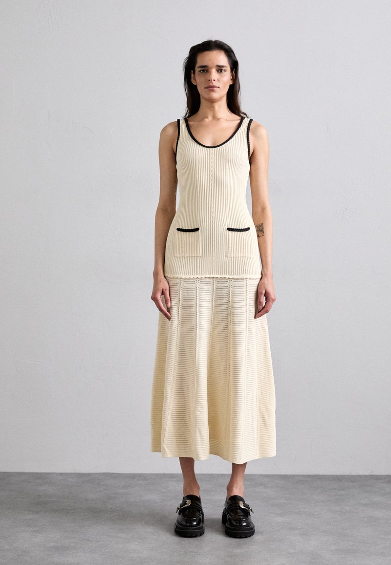 Vestido de tirantes de punto acanalado en crema con bordes negros y dos bolsillos frontales. Falda suelta de longitud hasta los tobillos con diseño texturizado. Zapatos de plataforma negros.