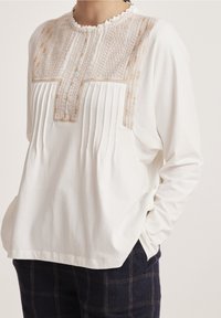 Top blanco de manga larga con frente plisado y canesú estampado beige. Presenta un cuello con volantes y un ajuste relajado. Combinado con pantalones oscuros a cuadros.